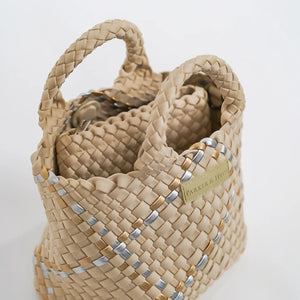 A woman with long brown hair, in a sleeveless cream knit top and blue jeans, holds a PARKER AND HYDE - MINI WOVEN TOTE BAG. The hand-woven tote by PARKER & HYDE features a subtle checkered pattern with light blue accents.