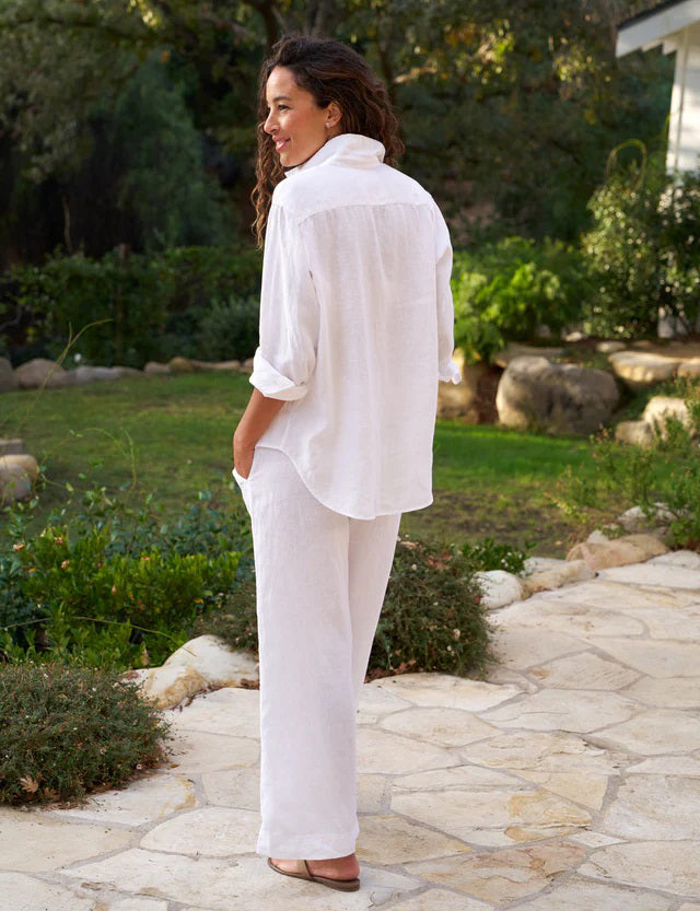 A person with long, wavy hair stands outdoors on a stone path, facing away from the camera. They are wearing a loose-fitting white shirt and wide-leg silhouette trousers from the Maisie Wide Leg Linen Pull On Pant for the Tuscany Getaway Set by Frank &amp; Eileen, hands in pockets. Lush greenery and trees are visible in the background, suggesting a garden or park setting.
