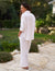 A woman with long hair is standing on a stone pathway outside, facing away from the camera. She is wearing the FRANK & EILEEN Eileen Relaxed Button-Up Shirt in Linen from the Tuscany Getaway Set paired with Maisie pull-on pants, both in a white, loose-fitting style. Her sleeves are rolled up and her hands are in her pockets. In the background, lush greenery and rocks reminiscent of a Tuscany Getaway set the scene.