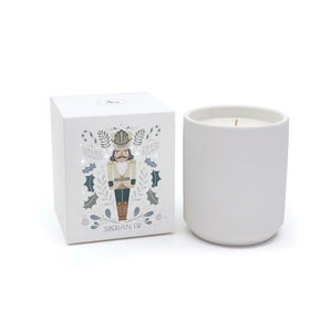 A NUTCRACKER SIBERIAN FIR CANDLE by ZODAX, housed in a minimalist white holder, is placed next to a decorative box featuring nutcracker art. Both items are elegantly arranged on a glittery tray, creating an enchanting atmosphere with fragrance notes that evoke cozy memories.