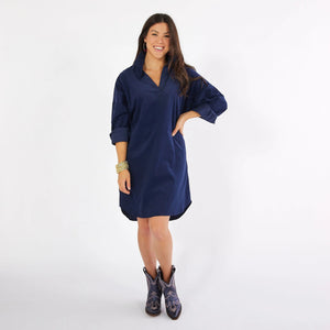 A person stands facing away, showcasing a contemporary chic style in the CARYN LAWN PREPPY Corduroy Dress. The dress is designed in a navy-blue hue and features hand-sewn green star patches on the sleeves. Their long hair cascades down to cowboy boots decorated with a star pattern, all set against a simple white studio backdrop that highlights the outfit's stunning details.