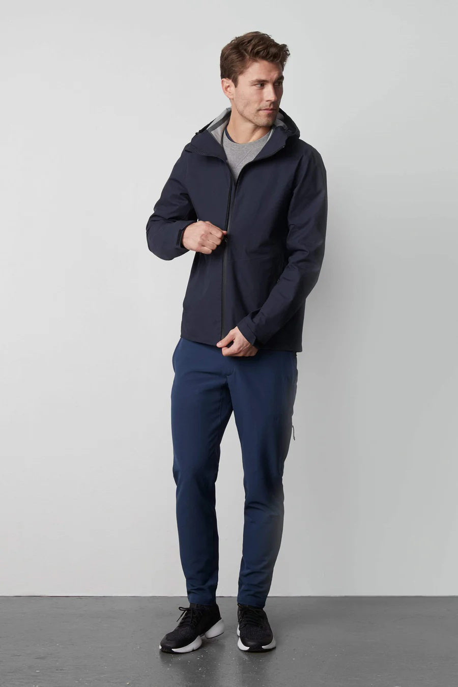 A man stands in front of a white wall wearing the MACK WELDON STORMCHASER JACKET with adjustable hood, paired with a gray shirt, blue pants, and black sneakers. He looks to the side while holding the jacket&#39;s zipper.