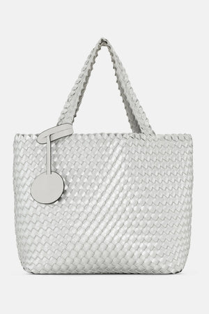 The WOVEN REVERSIBLE TOTE BAG by LINES OF DENMARK is a white tote featuring two shoulder straps and a circular faux-leather tag on one strap. This shopper showcases a textured, interlaced pattern and reversible design, providing stylish and elegant dual styling options. The image background is plain white.