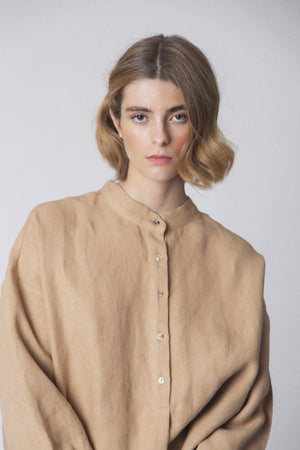 A person with short, wavy blonde hair wears the LOESS - FLORENCE LINEN BLOUSE in beige. Featuring gathered fabric detail on the back, it stands out against a neutral light gray background, showcasing timeless style and sustainable fashion by LOESS.