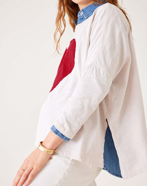 A person with long hair sits on a wooden stool against a white background, wearing an oversized MER SEA Catalina Long Sleeve Slub Tee with Heart, white pants, and blue boat shoes. They look at the camera and smile.