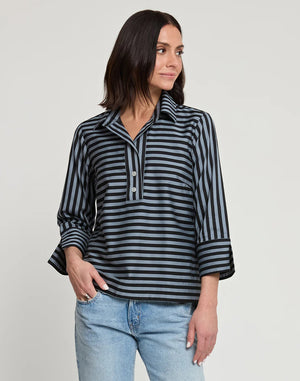 A woman with shoulder-length dark hair wears the HINSON WU - AILEEN 3/4 SLEEVE STRIPE GINGHAM COMBO SHIRT, a black and beige striped blouse featuring a small collar and a partial zip at the front. She pairs it with black pants and stands against a plain white background, one hand in her pocket.