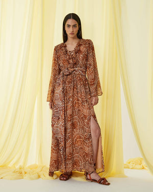 The SFIZIO - RUST PRINT RUFFLED DRESS by SFIZIO is a long, full-sleeved, flowy dress made in Italy, featuring a brown and beige spiral pattern. It boasts a V-neckline, ruffled detailing on the bodice, and an elastic waistband. Crafted from lightweight viscose creponne, it’s perfect for warm weather.