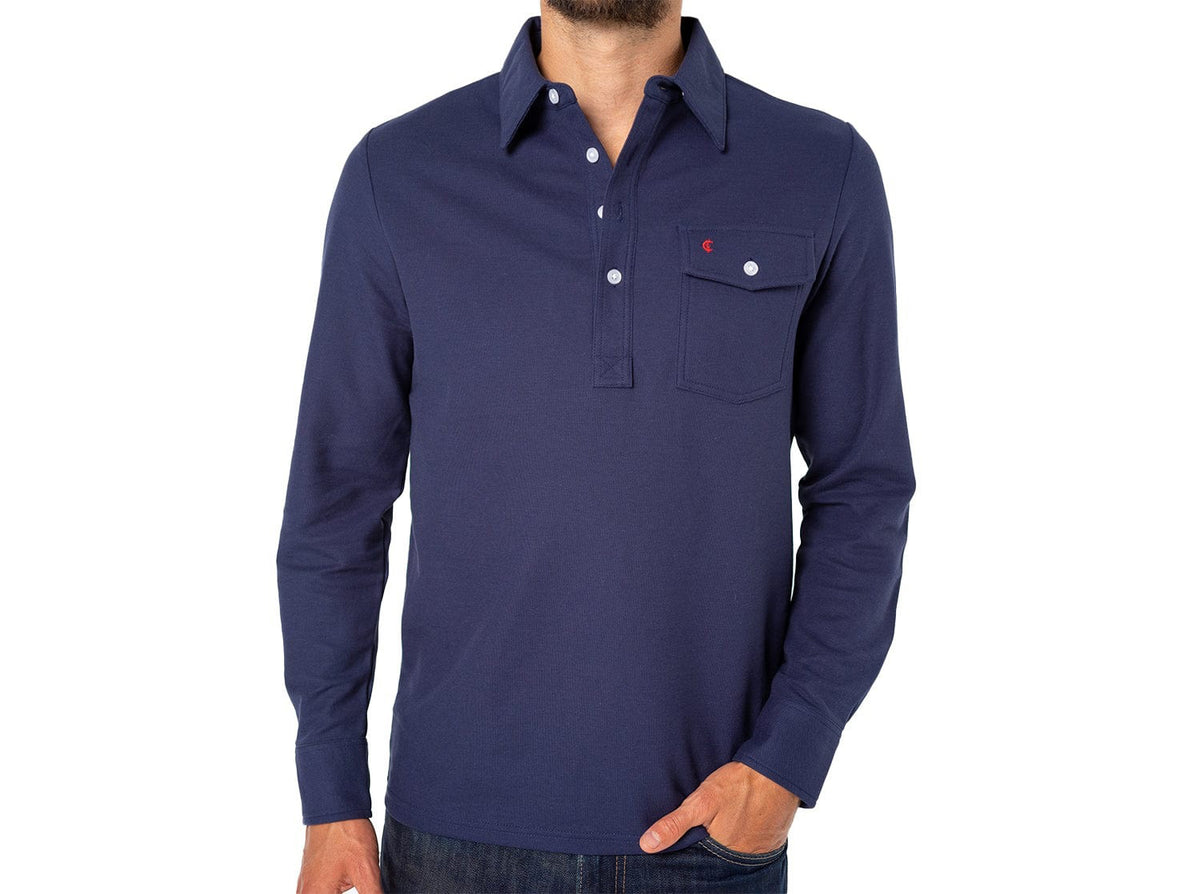 A man wears the CRIQUET Long Sleeve Players Polo Shirt in Peacoat, made from soft Peruvian Pima cotton with a buttoned collar and chest pocket. He stands with one hand in his jeans pocket against a white background.