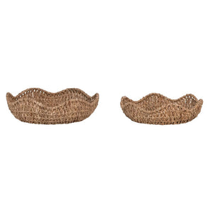 A wicker basket lined with parchment paper contains several dark purple tomatoes, positioned on a light-colored surface next to another empty "BRAIDED BANKUAN BOWL WITH SCALLOPED EDGE (SMALL)" by CREATIVE COOP and a piece of paper. The arrangement evokes a rustic or homey atmosphere.