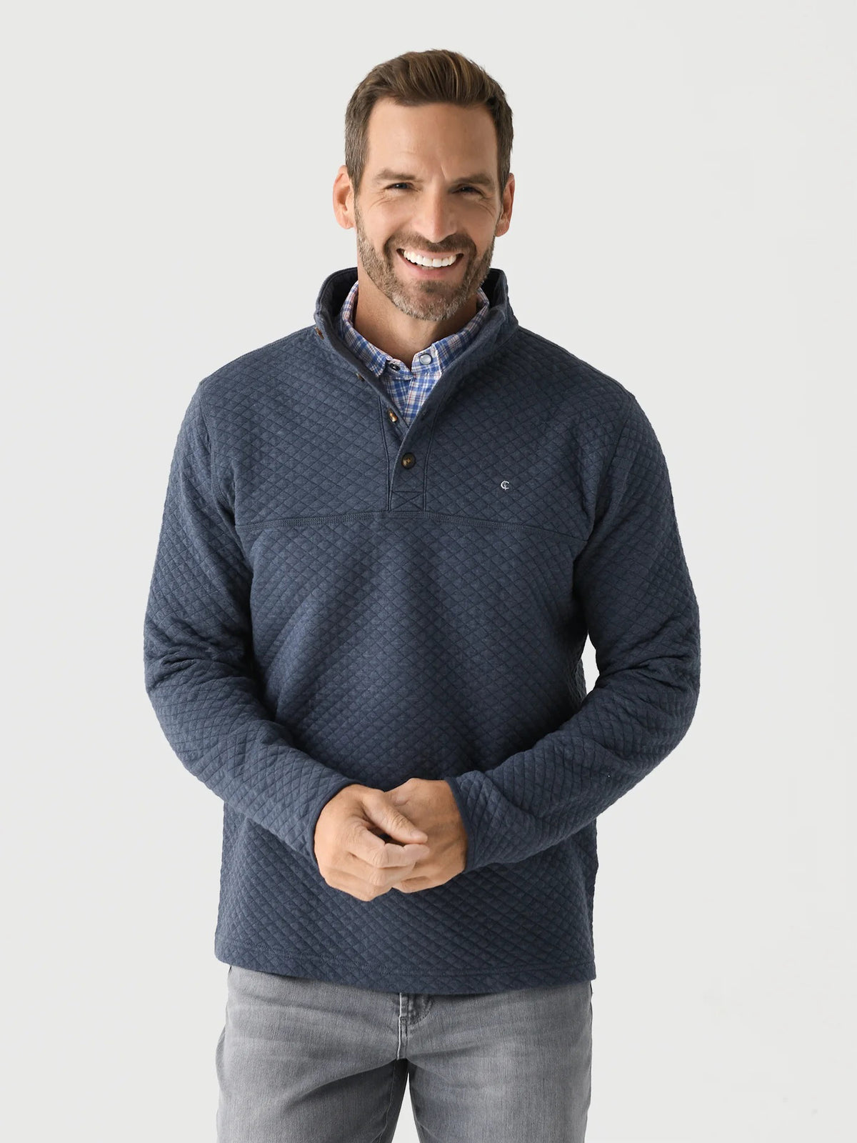 A man with short brown hair and a beard smiles while wearing the CRIQUET - QUILTED PULLOVER IN HEATHER NAVY over a blue plaid shirt and light gray jeans, standing against a plain white background.
