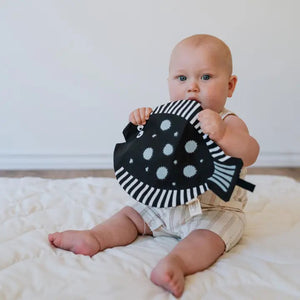 The WEE GALLERY - ORGANIC CRINKLE FISH by WEE GALLERY is a charming black, fish-shaped oven mitt made from organic cotton, featuring white polka dots scattered across its surface. It includes a handle resembling a fish tail and an opening at the side for inserting your hand. Additionally, there's a black tag attached near the middle.