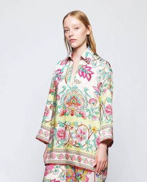 A person with long blonde hair stands against a plain background, wearing the MIRTO 1956 - FLUID BLOUSE MULTICOLOR FLORAL by MIRTO 1956. The uplifting blouse features a vibrant, floral pattern in pink, green, yellow, and blue. The 3/4 sleeves and V neckline enhance the intricate designs, creating a bright and stylish look.