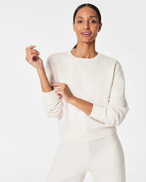 A woman stands confidently against a plain white background, wearing the SPANX Aireessentials Crew Neck Pullover and matching sweatpants made from luxurious comfort spacer fabric. She has her hair pulled back and her left hand is raised slightly. She smiles gently with her lips closed.