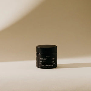 A close-up shot of an open jar filled with PRESTON - CREAM POMADE UNSCENTED. The smooth, frizz-free texture of the pomade is visible within the dark-colored container. The jar is placed on its side against a neutral background.