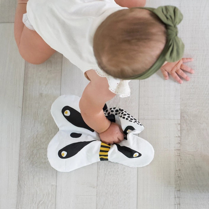 The WEE GALLERY - BUTTERFLY ORGANIC CRINKLE is a children&#39;s soft fabric toy, crafted from organic cotton. It features a butterfly shape with a smiling face in the center and a striped yellow and black body. The white wings are adorned with black patterns and yellow accents, providing visual stimulation. Additionally, it includes a small loop at the top for hanging.