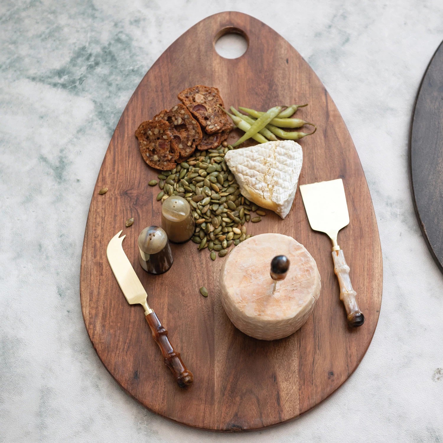 The ACACIA CHEESE BOARD WITH HANDLE by CREATIVE COOP is crafted from elegant acacia wood, featuring an oval shape with a small hole near the top. The natural grain pattern and smooth finish make it perfect for both serving cheese and general kitchen prep.