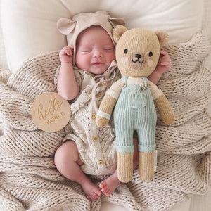 Two CUDDLE & KIND Charlie the Honey Bears are shown standing side by side. Both handmade bears are wearing light blue overalls and have beige fur with cute embroidered facial features. The larger 20" bear is on the left, and the smaller bear on the right. These adorable hand-knit dolls make perfect baby shower gifts, set against a plain white background.