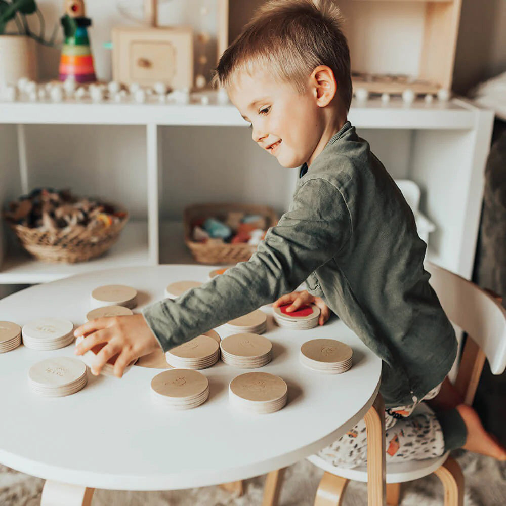 A young boy in a green shirt plays intently with the LITTLE MATCHABLES GAME SET by LILY & RIVER on a white table in a Montessori playroom. The room is adorned with various toys and baskets, and the boy appears focused on enhancing his fine motor skills by arranging and stacking the round wooden discs as part of this engaging memory game.