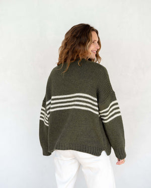 A person with long, wavy brown hair is wearing the MERSEA - SEACOAST STRIPE SWEATER, a loose-fitting, cozy blue knit sweater with white stripes on the sleeves, paired with black pants. They are standing against a plain, light-colored background and facing left, with their hands near their face.