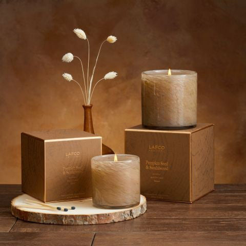 A candle in a transparent glass container, featuring a rich brown hue, is placed beside a coordinating brown box adorned with gold details. The set exudes a gourmand aroma and is elegantly labeled as "LAFCO - Pumpkin Seed & Sandalwood Candle 6.5oz." The candle's texture appears soft like fur.