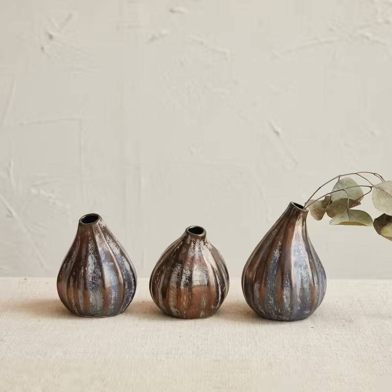 Three STONEWARE FIG SHAPED VASES by CREATIVE COOP in rustic, earthy tones are arranged on a neutral surface. Each vase features a reactive glaze finish, with one displaying a sprig of dried leaves. The scene is set against a subtly textured wall in the background.