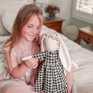 The ALIMROSE - SOFIA BUNNY BLACK CHECK is a handcrafted, ethically stuffed doll from ALIMROSE. It wears a black and white gingham dress with a matching fabric headband that accentuates its long ears, and it carries a small bag around its neck. This charming bunny, made from the finest materials, features simple stitched facial details.