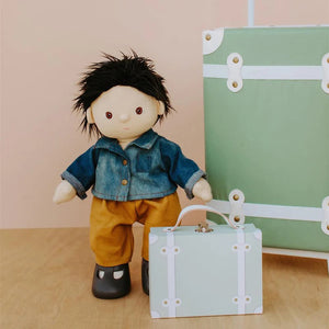 A Dinkum Doll from Olli Ella USA's Travel Togs collection, featuring dark hair and dressed in a denim jacket, mustard pants, and black shoes, stands on a wooden surface. Beside the doll is a teal See-Ya Suitcase with white trim and hardware. In the background, another larger teal suitcase is partially visible.