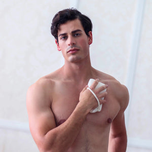 A muscular man with dark hair stands shirtless against a light background. He holds a BODY SCRUBBER by DAILY CONCEPTS, its gentle exfoliating texture pressed to his chest as he appears to be gently patting or wiping his skin. His facial expression is neutral.