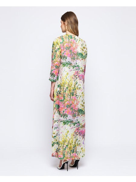 A person stands with their back turned, wearing the MIRTO 1956's Linen Brush Stroke Print Dress featuring floral watercolor patterns in pink, green, and yellow. They have long brown hair and are donning black high heels against a plain white background.