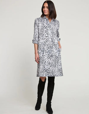 A woman with shoulder-length dark hair is wearing a knee-length HINSON WU - CHARLOTTE 3/4 Sleeve Snow Leopard Sateen Dress. She is also sporting black knee-high boots and has her hands in the dress pockets. The background is plain white.