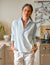 A person stands in a kitchen, hands in pockets, wearing the FRANK & EILEEN PATRICK POPOVER HENLEY IN ICE and white pants, with a stove and kettle behind them. The marbled backsplash displays greenery and a decorative dish on the countertop.