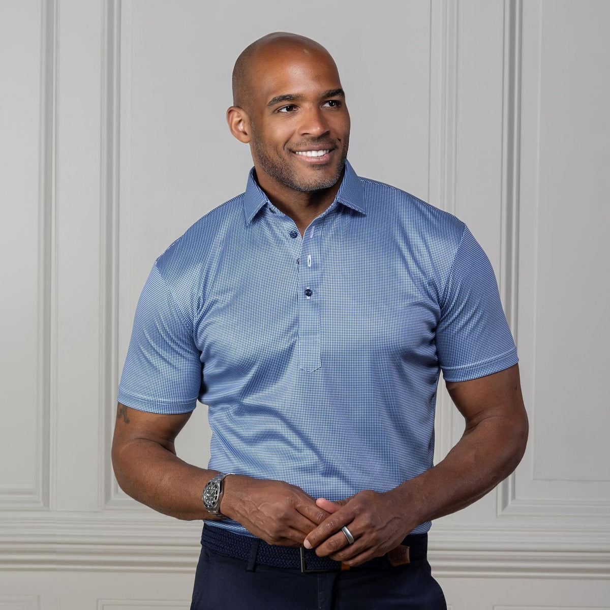 A man in sunglasses and a COLLARS AND CO - Semi Spread Collar Polo in Navy Houndstooth stands outdoors by a stone building and green plants, gazing to the side with one hand in his pocket.