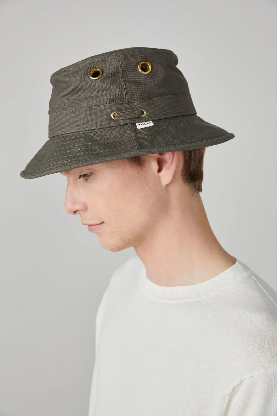 A young man in a white shirt is shown in profile wearing the TILLEY - ICONIC T1 BUCKET HAT in dark gray with golden eyelets. The UPF 50+ sun protection hat stands out against the plain, light gray background.