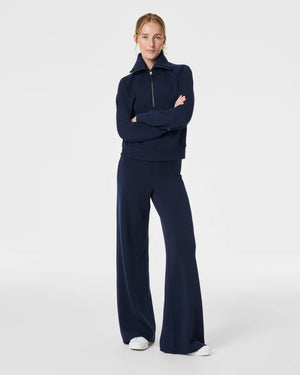 A person stands against a plain background wearing a matching two-piece outfit. The ultimate throw-on-and-go style consists of a long-sleeve, quarter-zip, high-collar pullover and SPANX AIRESSENTIALS WIDE LEG PANT in a deep blue color. They also wear white sneakers and have their hair pulled back.