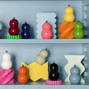 A display of ADDISON ROSS LONDON - CHUBBIE SALT OR PEPPER MILLS in various vibrant shades, including red, blue, pink, orange, white, black, and gray. They are arranged on shelves alongside other salt mills and pepper grinders boasting ceramic mechanisms amidst colorful geometric shapes against a pastel blue backdrop.