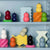 A display of ADDISON ROSS LONDON - CHUBBIE SALT OR PEPPER MILLS in various vibrant shades, including red, blue, pink, orange, white, black, and gray. They are arranged on shelves alongside other salt mills and pepper grinders boasting ceramic mechanisms amidst colorful geometric shapes against a pastel blue backdrop.