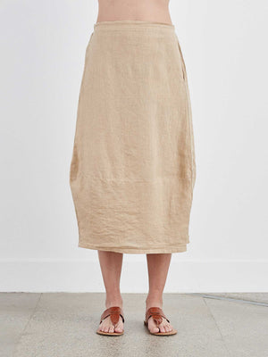 The CUT LOOSE - SIDE PLEAT SKIRT by CUT LOOSE hangs on a hanger against a plain grey background. The long white maxi skirt appears to be made of lightweight, slightly wrinkled linen fabric, with a simple design free of patterns or embellishments.