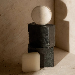 A minimalist arrangement of geometric shapes, featuring a white sphere perched on two black rectangular blocks stacked vertically, with a white cube placed at the base. The objects cast soft shadows against a textured wall, reminiscent of the simplicity seen in PRESTON's premium bar soap packaging designs.