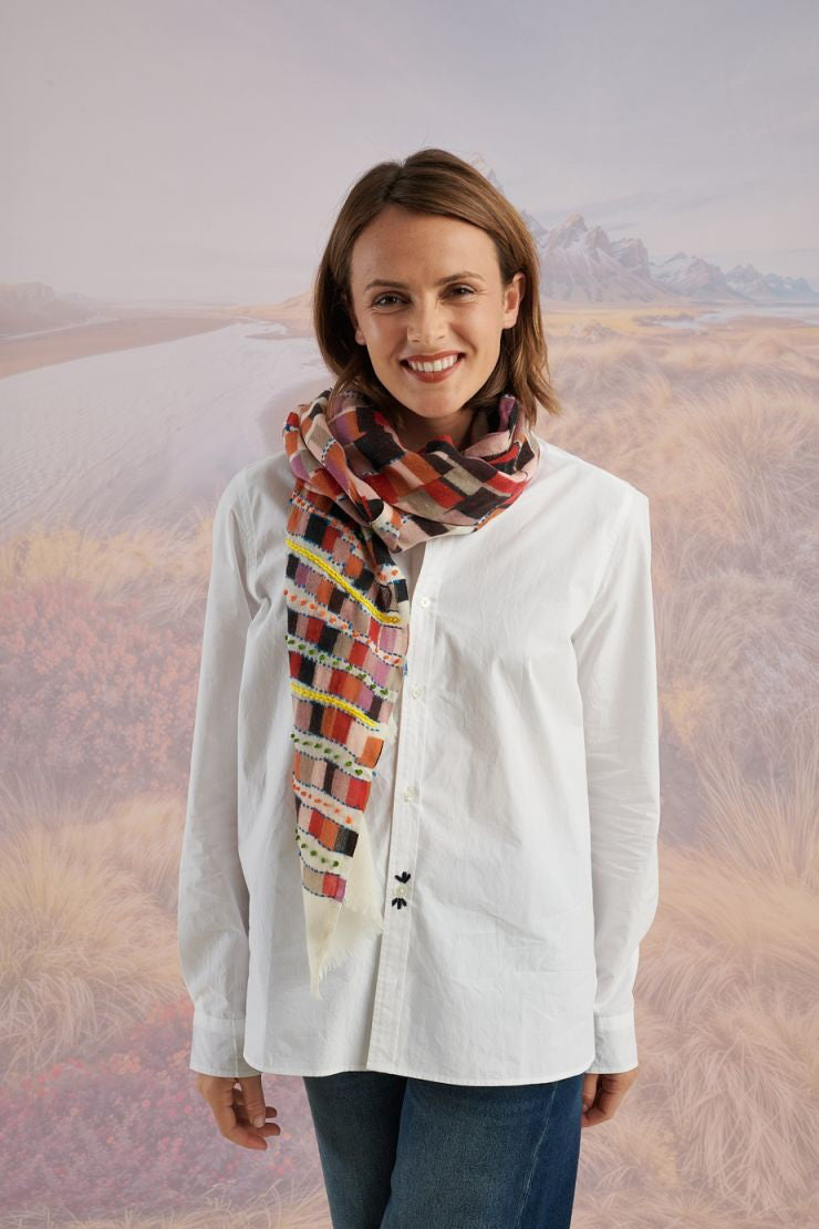 A woman with light brown hair wears a cream shirt and the STORIATIPIC - ANOUK WOOL SCARF, an elegant hand-embroidered accessory by STORIATIPIC, wrapped around her neck. Mountains and plains form a soft, scenic backdrop.