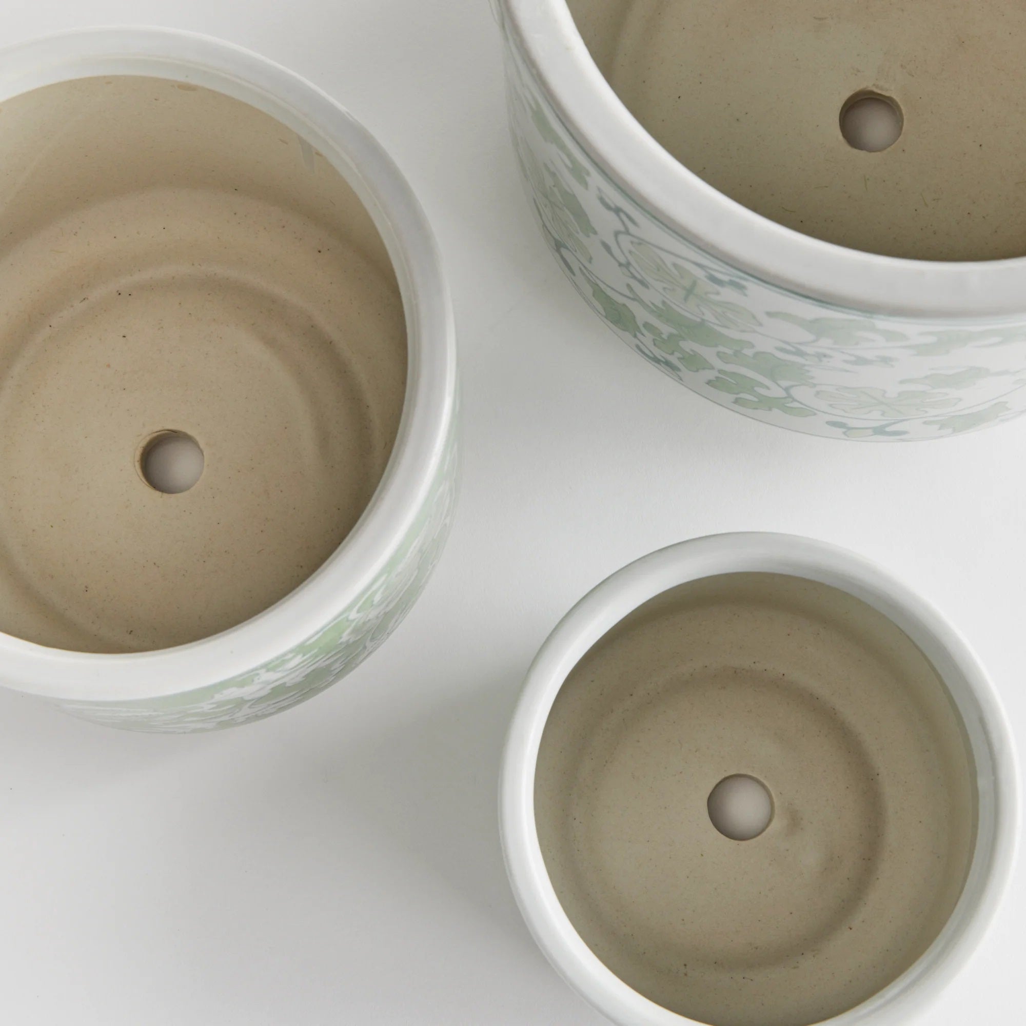 Three NAPA HOME AND GARDEN - IMPERIAL CYLINDER POTs with drainage holes are arranged on a white surface. These handcrafted pieces feature subtle green chinoiserie patterns, varying in size and viewed from above to showcase their unique elegance.