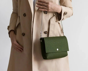 A person in an olive green trench coat holds the WOOLBERRY WOOL FELT CROSSBODY BAG in light gray with a black strap; only their torso and hand are visible.