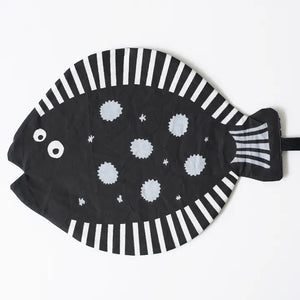 The WEE GALLERY - ORGANIC CRINKLE FISH by WEE GALLERY is a charming black, fish-shaped oven mitt made from organic cotton, featuring white polka dots scattered across its surface. It includes a handle resembling a fish tail and an opening at the side for inserting your hand. Additionally, there's a black tag attached near the middle.