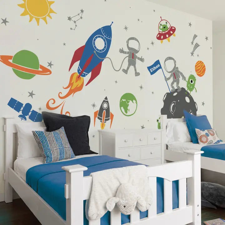 Children&#39;s bedroom with two white beds featuring blue and striped bedding. The wall is decorated with SIMPLE SHAPES - OUTER SPACE REMOVABLE WALL DECALS by SIMPLE SHAPES, including rockets, astronauts, planets, stars, and aliens. The wall also has a constellation and sun illustration.