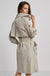 A woman with long brown hair poses in the ADROITE ATELIER - RAPHAEL TRENCH COAT by ADROIT ATELIER. This light beige trench coat highlights rolled-up sleeve details, a belted waist, and a flap on the back. She stands against a plain gray background, glancing over her shoulder.