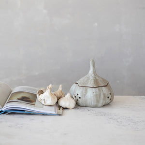 The STONEWARE GARLIC STORAGE by CREATIVE COOP is a light gray container with a speckled, reactive glaze, featuring a rounded base, tall tapered lid with stem, and side holes. Its rustic finish is perfect for stylishly storing garlic or onions.