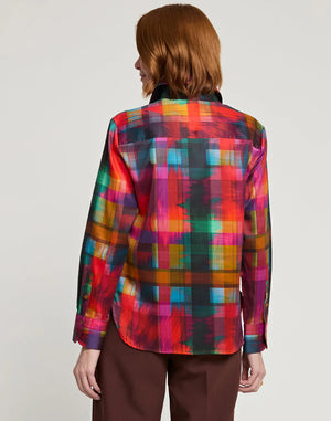 A person with shoulder-length auburn hair is shown from the back, wearing the HINSON WU - Margot Long Sleeve Satin Shirt in Festive Plaid. The multicolored plaid shirt features a collar and includes shades of red, green, yellow, and pink. It is paired with brown pants.