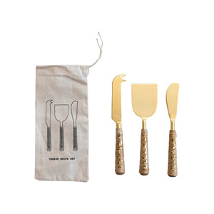 The CREATIVE COOP STAINLESS STEEL CHEESE KNIFE WITH RATTAN HANDLES SET includes three knives with rattan-wrapped handles and stainless steel blades. These tools are accompanied by a cloth pouch adorned with an illustrated pattern of the knife set, all displayed on a textured off-white surface.