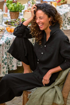 A woman with wavy blonde hair stands indoors in a loose black shirt and FRANK & EILEEN’s Maisie Wide Leg Pull On Pant in Washed Linen Black, barefoot on a wooden floor near a chair, vase of green flowers, and staircase.