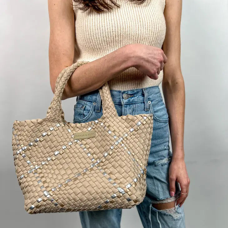 A person in a beige sleeveless top and ripped blue jeans holds a Parker & Hyde - Beige Metallic Woven Tote Bag, featuring a checkered design with subtle white accents and two handles. The background is plain light gray.