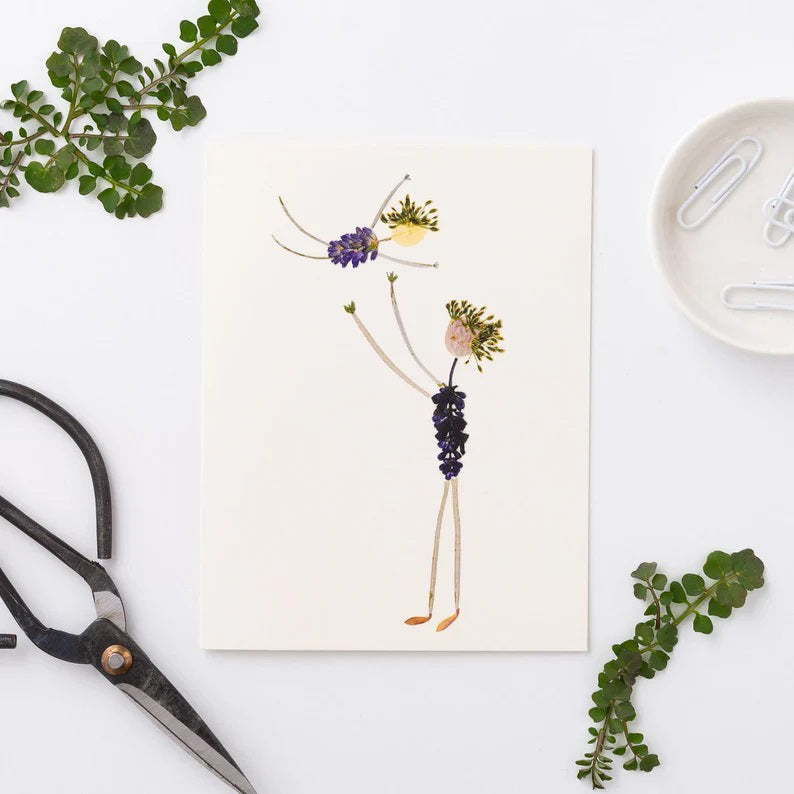 Two whimsical figures crafted from pressed flowers and leaves are playfully depicted, with one figure lifting the other above its head. These figures, featuring leafy limbs and floral bodies set against a white background, make an ideal choice for a Father's Day card. This card is crafted by Petal People Press on FSC-certified cardstock.